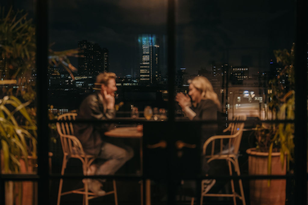 A couple seated at a rooftop restaurant in London, softly lit against a blurred city skyline at night.
