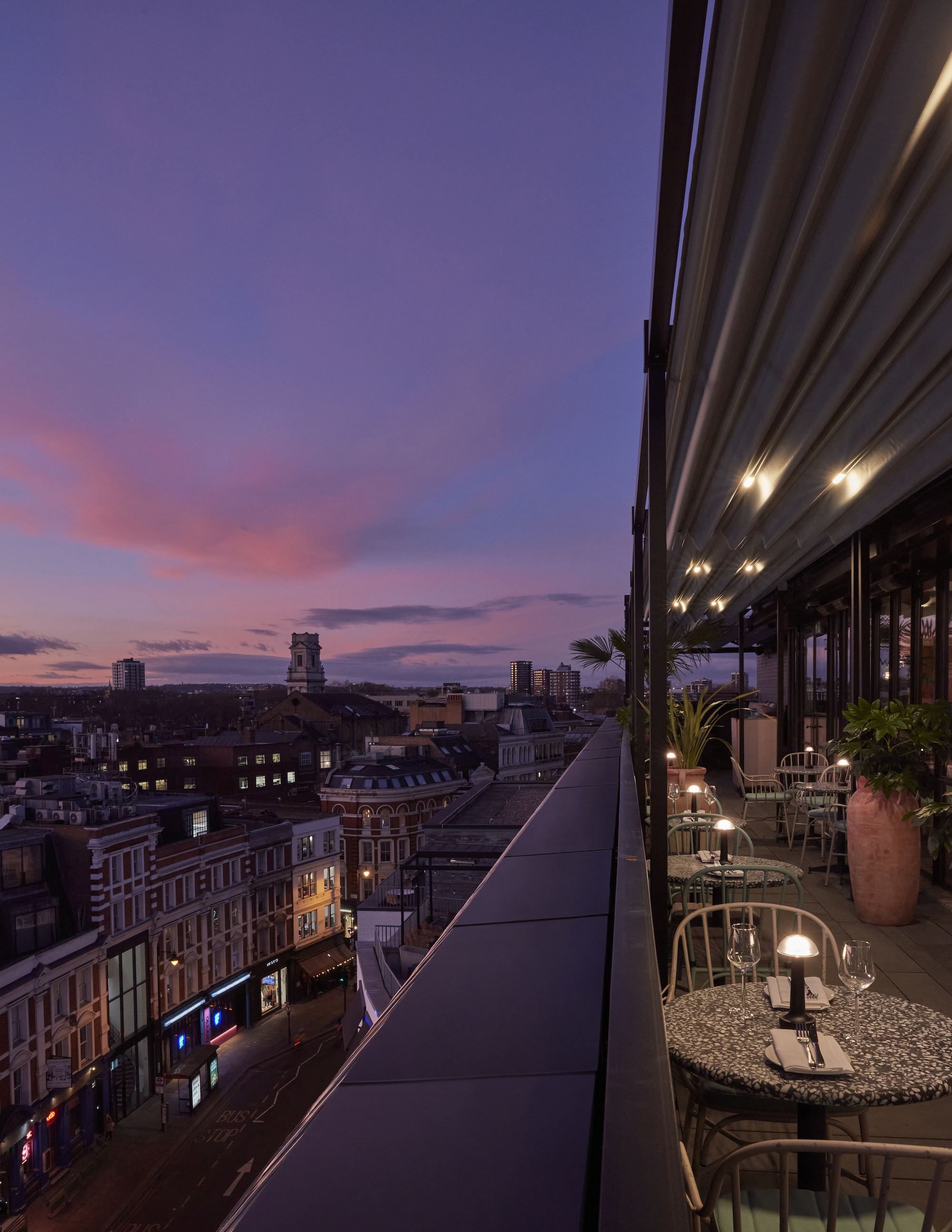 A rooftop terrace at One Hundred Shoreditch with twinkling lights, lush greenery, and a panoramic view of London at sunset.