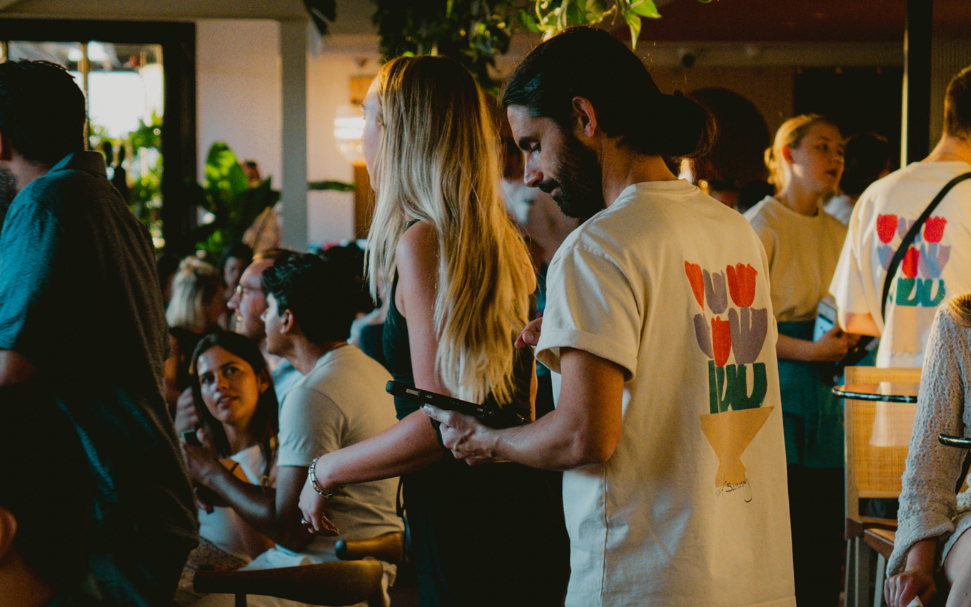 A lively bar scene where a male server in a floral-printed T-shirt in a dimly lit, stylish space filled with greenery.
