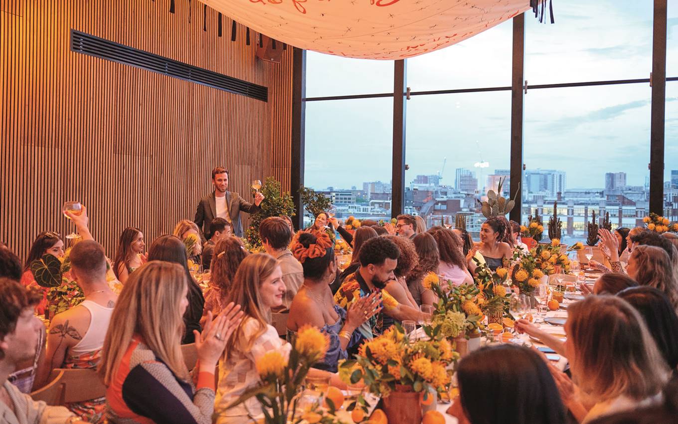 A lively dining event in London with guests seated at a long table adorned with bright yellow flowers and greenery.