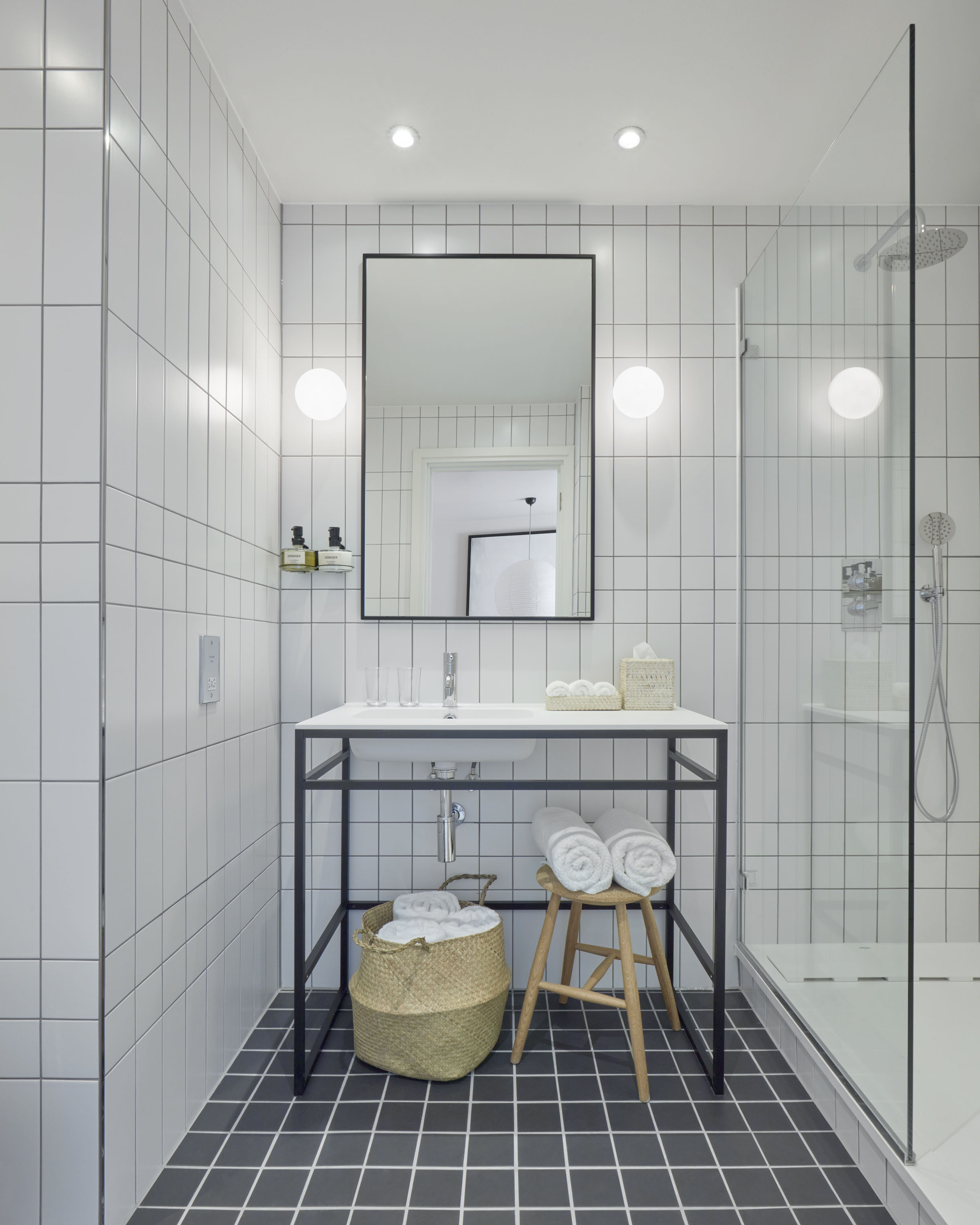 White-tiled hotel bathroom with a minimalist sink, rolled towels, and woven baskets on the black and white tiled floor.