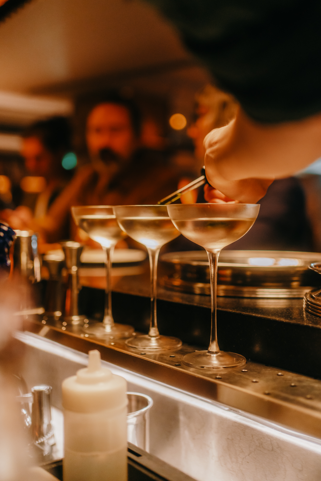 A bartender using tweezers to garnish three coupe glasses filled with clear cocktails in a dimly lit bar in London.