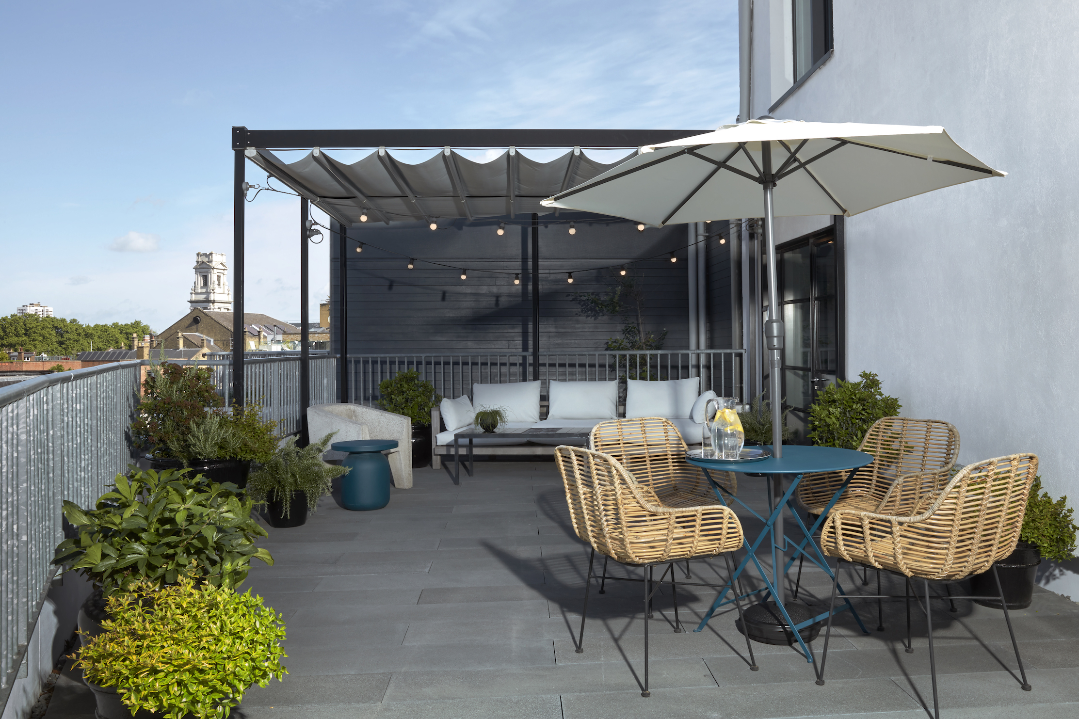 Cosy rooftop lounge area with rattan chairs at One Hundred Shoreditch with a blue table, string lights and London views.
