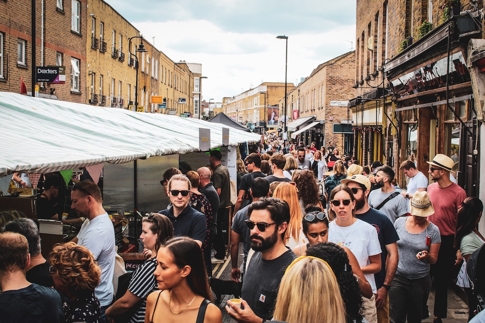 Crowded Broadway Market in London with people shopping and interacting near vendor stalls under striped canopies.