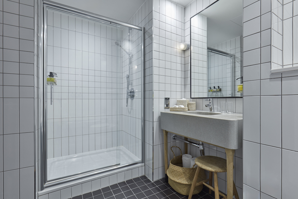 Tiled hotel bathroom at One Hundred Shoreditch with glass shower, vanity, and storage baskets.