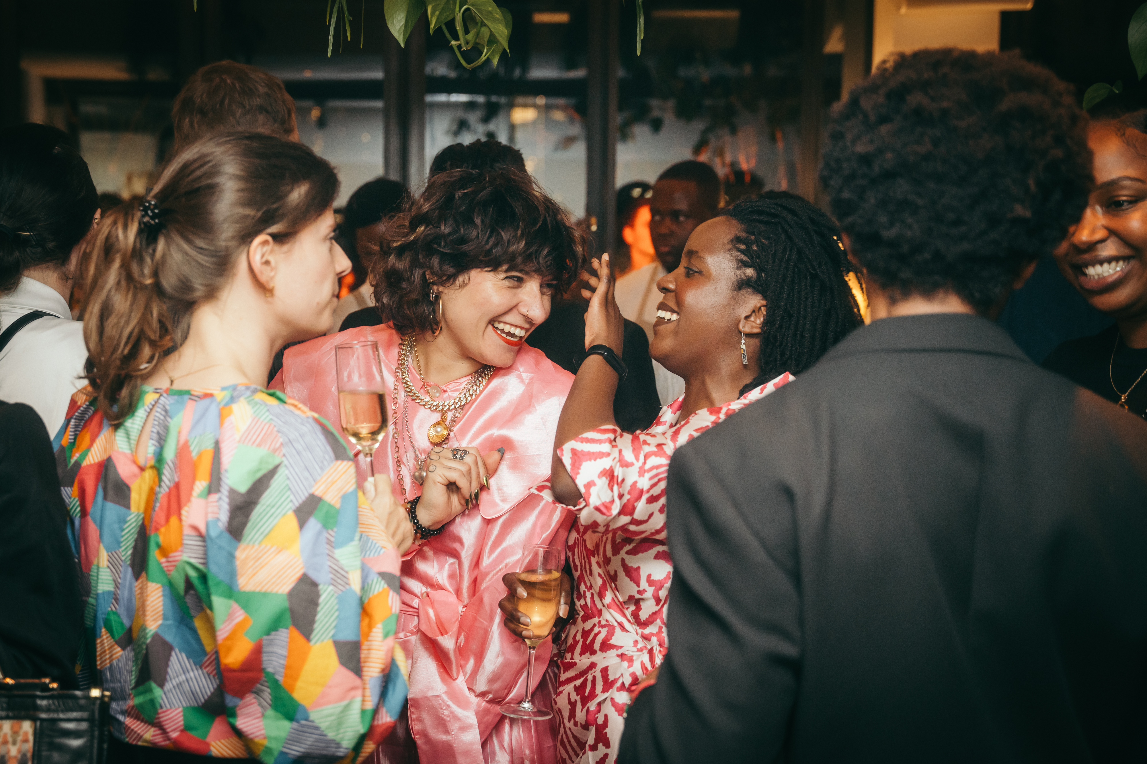 Friends at a central London party, chatting and laughing while holding champagne glasses, dressed in vibrant clothing.
