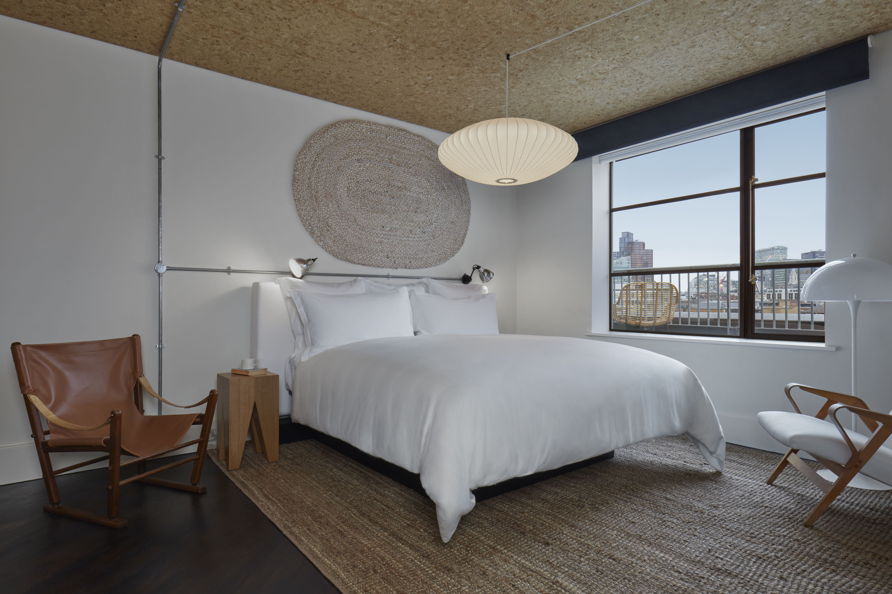 A cosy hotel bedroom with a white bed, leather lounge chair, soft lighting, and a large window with a city view.