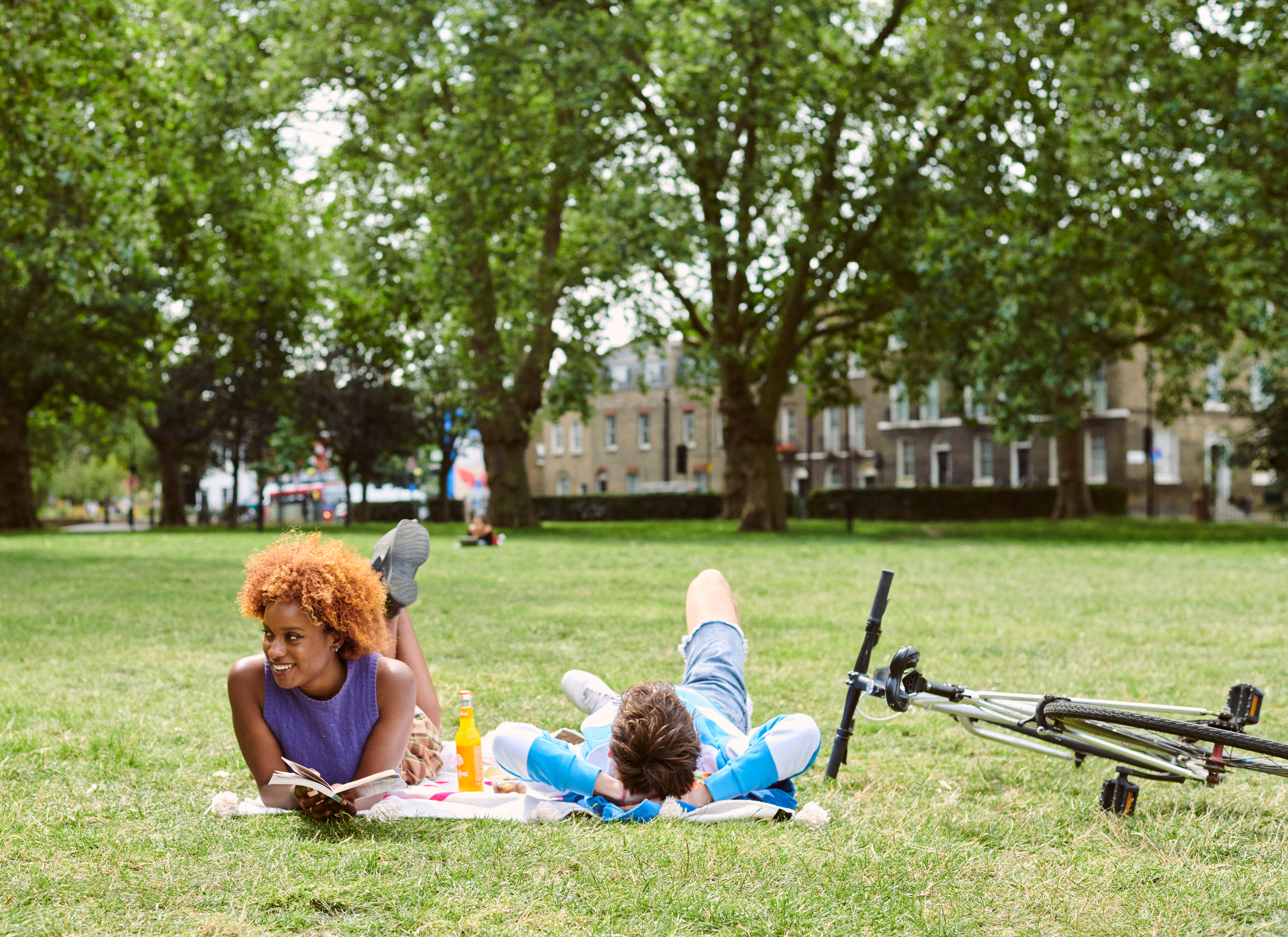 Two friends relaxing on a blanket in a London park, reading and chatting, with a bicycle nearby and trees surrounding them.