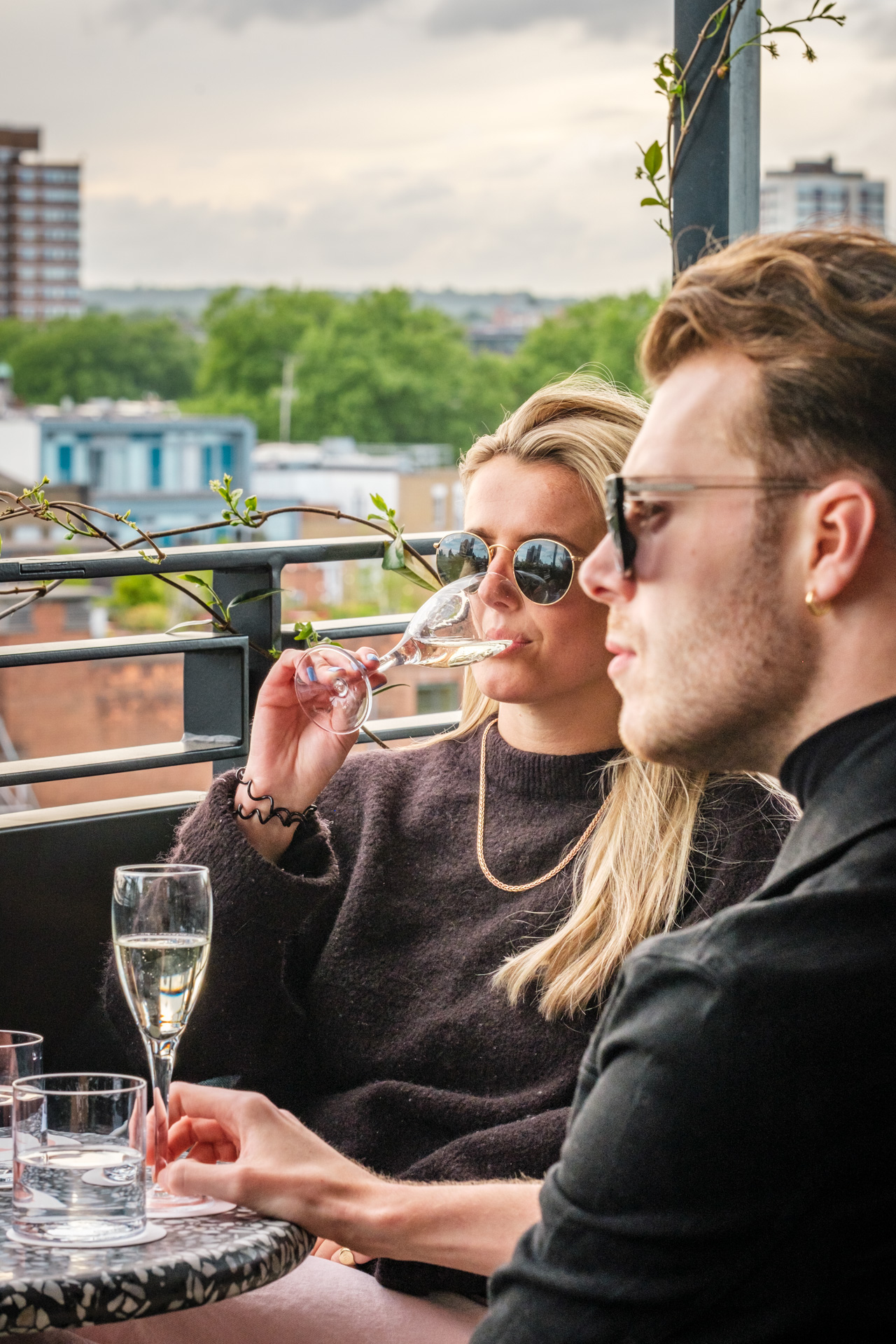 A woman sipping champagne outdoors with a man beside her, enjoying a drink on a London hotel’s rooftop terrace.