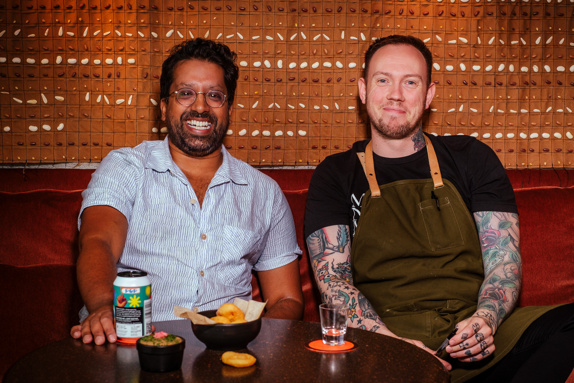 Mr Lyan and Alex smiling while seated at the Seed Library bar in London with drinks and snacks in a warmly lit setting.