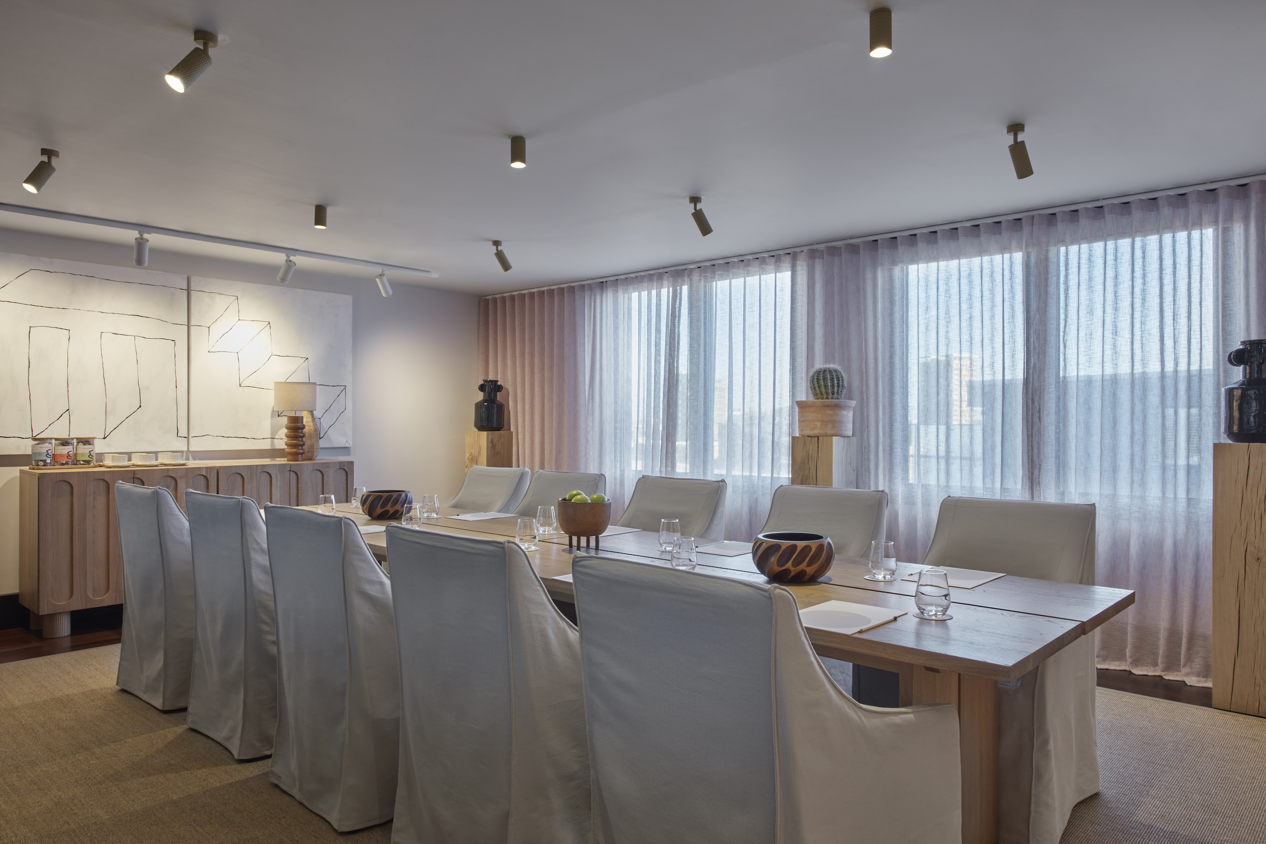 A modern meeting room in London with a large wooden table, white slipcovered chairs, abstract wall art, and natural light.