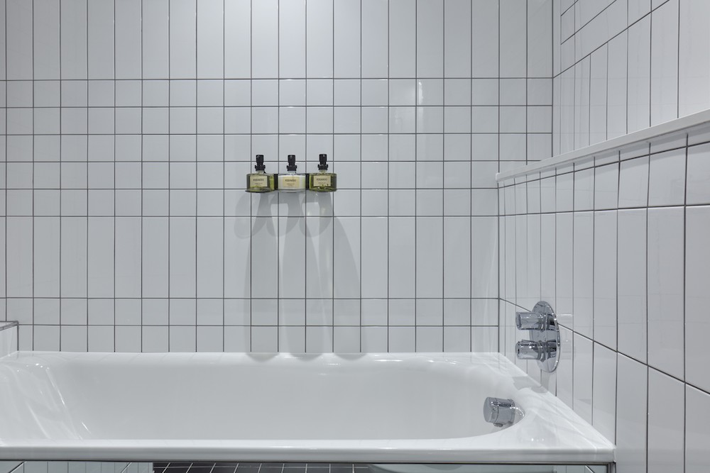 Modern white tiled bathtub with mounted bath products against the wall in a hotel room at One Hundred Shoreditch in London.