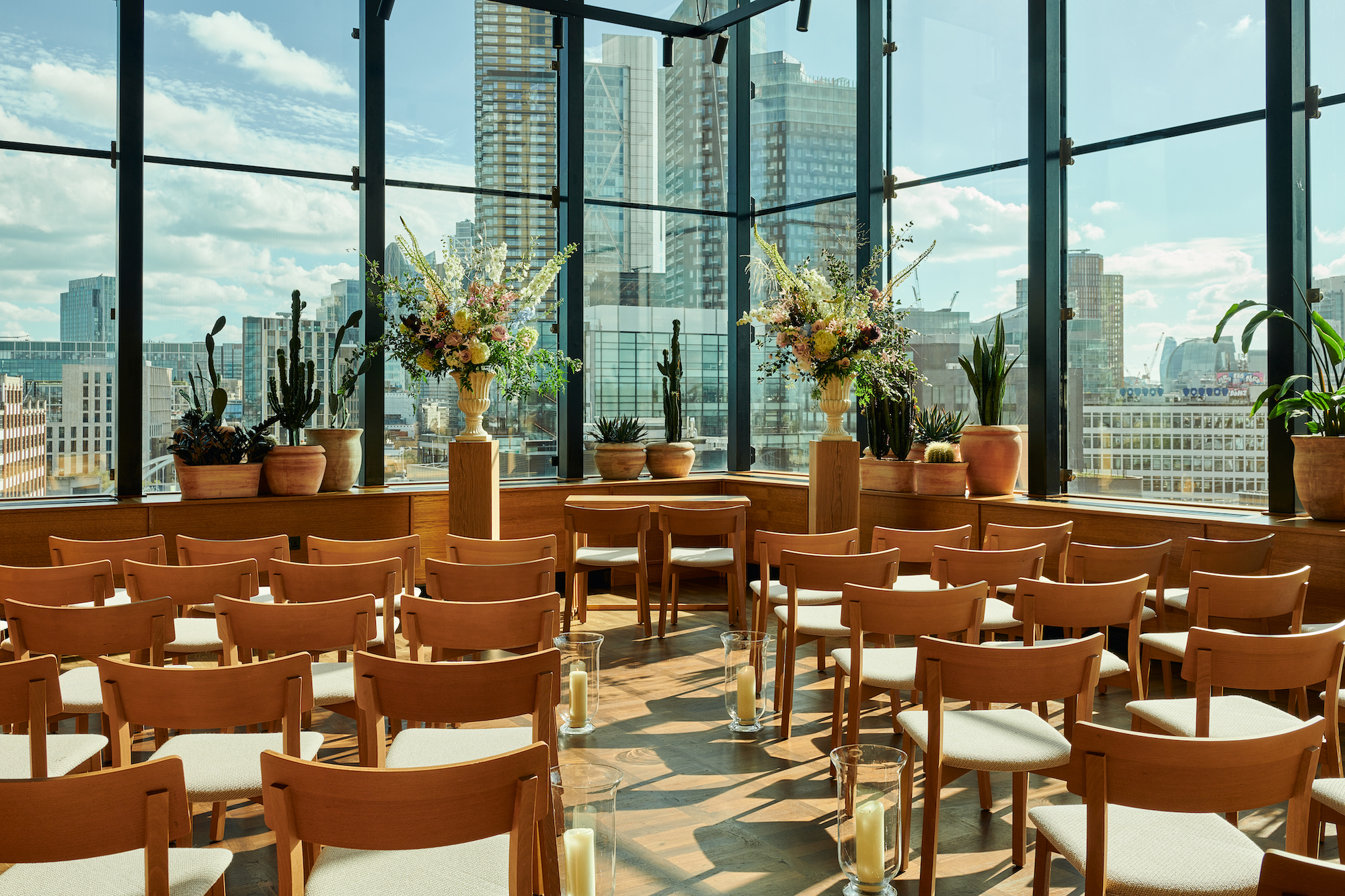 Sunlit room set for an event in London at One Hundred Shoreditch with wooden chairs, flowers, and city views through windows.