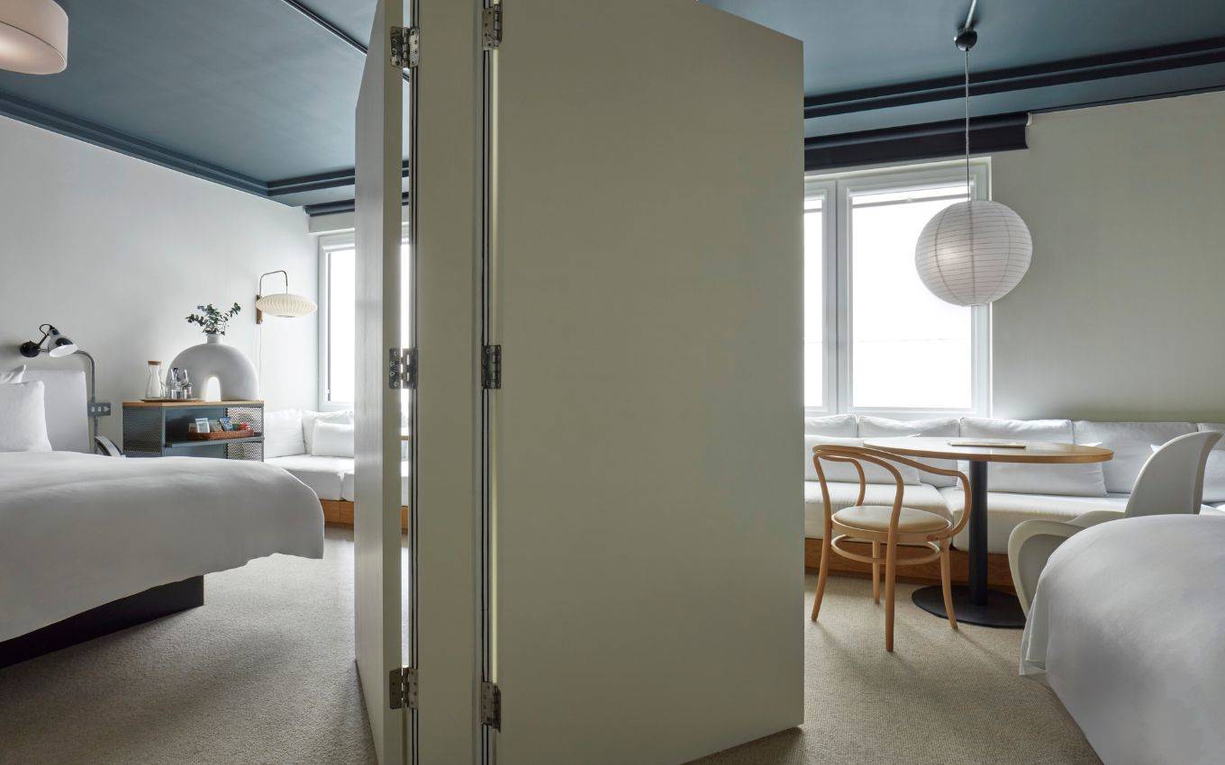 A spacious hotel room in London with a foldable partition, two beds, a seating area, and large windows letting in natural light.