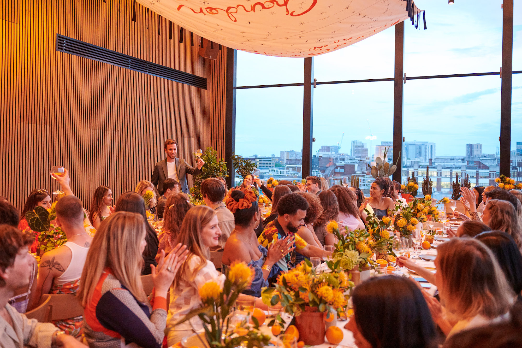 An event space in London with long tables decorated with yellow flowers, filled with guests enjoying a city view.