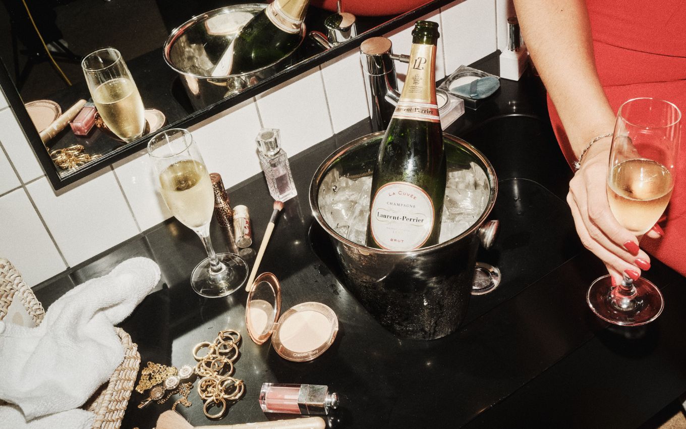 A bottle of Laurent-Perrier in an ice bucket sits on a vanity surrounded by makeup and accessories.