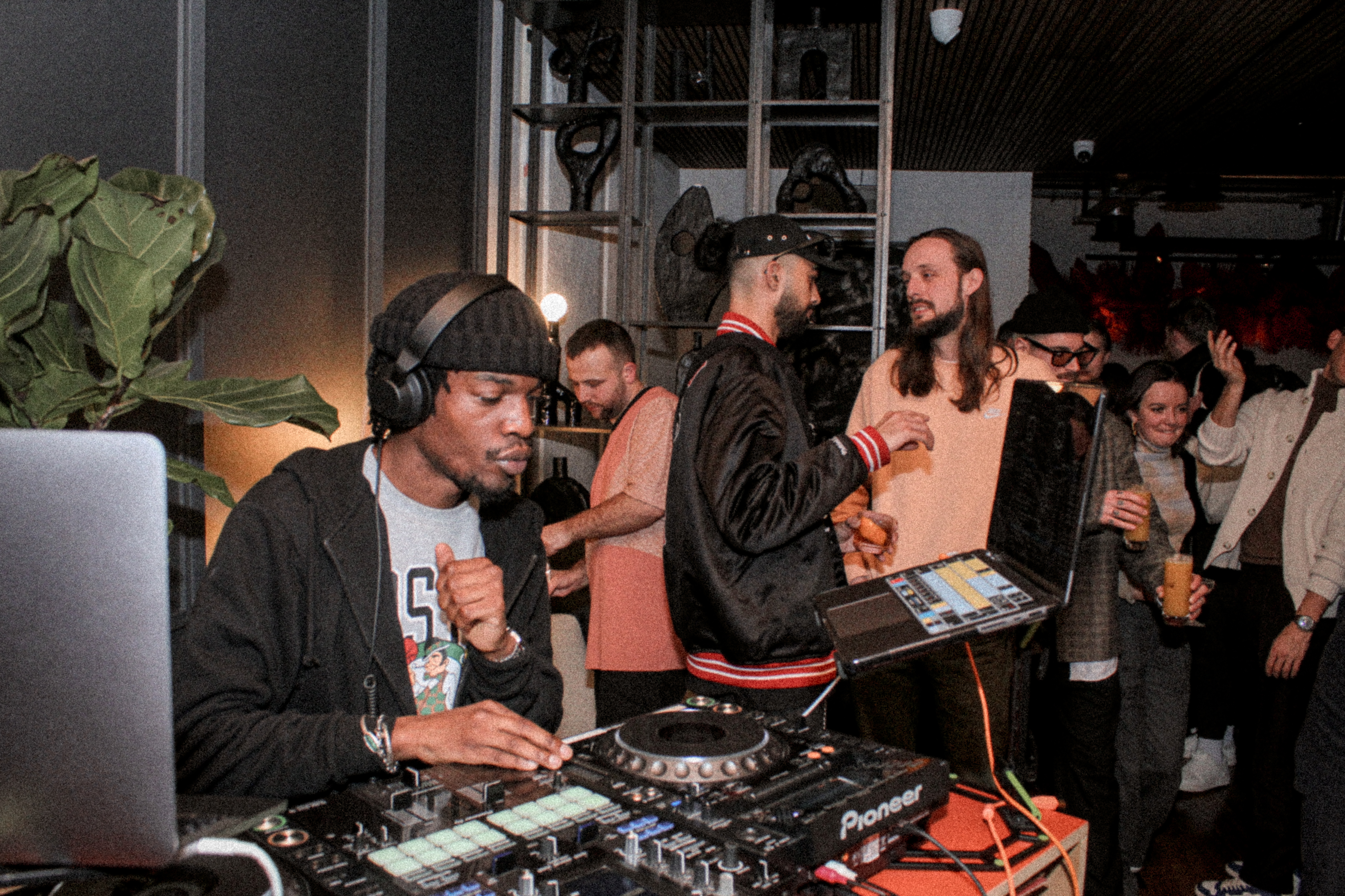 A DJ in a black beanie and headphones playing a set at One Hundred Shoreditch, while guests chat and dance nearby.