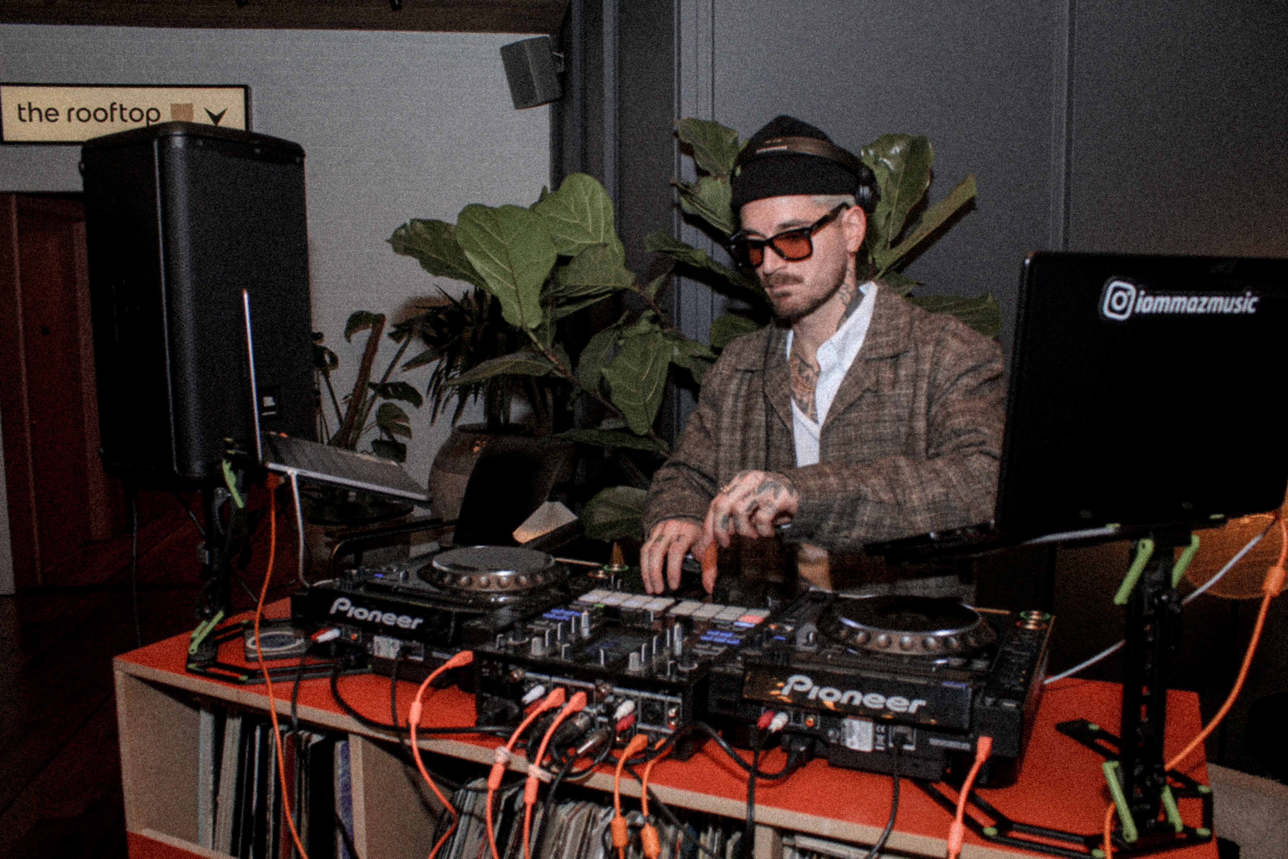 A DJ in sunglasses and a plaid jacket performing at a London bar, with Pioneer equipment and plants in the background.