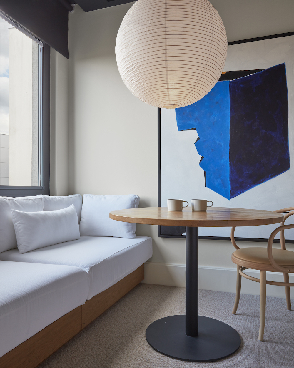 Cosy hotel room corner with a round wooden table, two cups, and a large blue abstract painting beside a soft white bench.