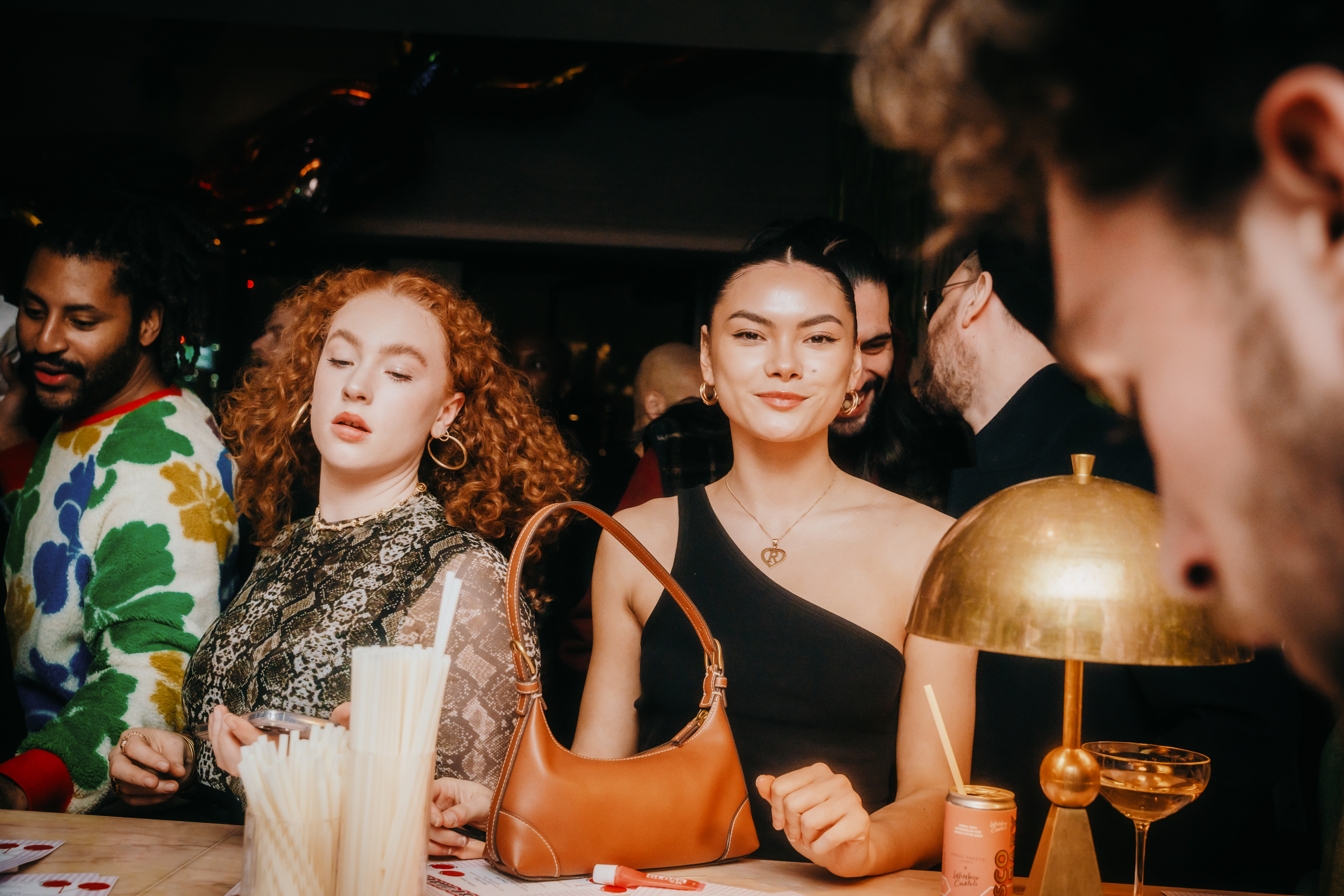 Guests socializing at a London bar with drinks, a warm lamp, and festive expressions enhancing the lively mood.