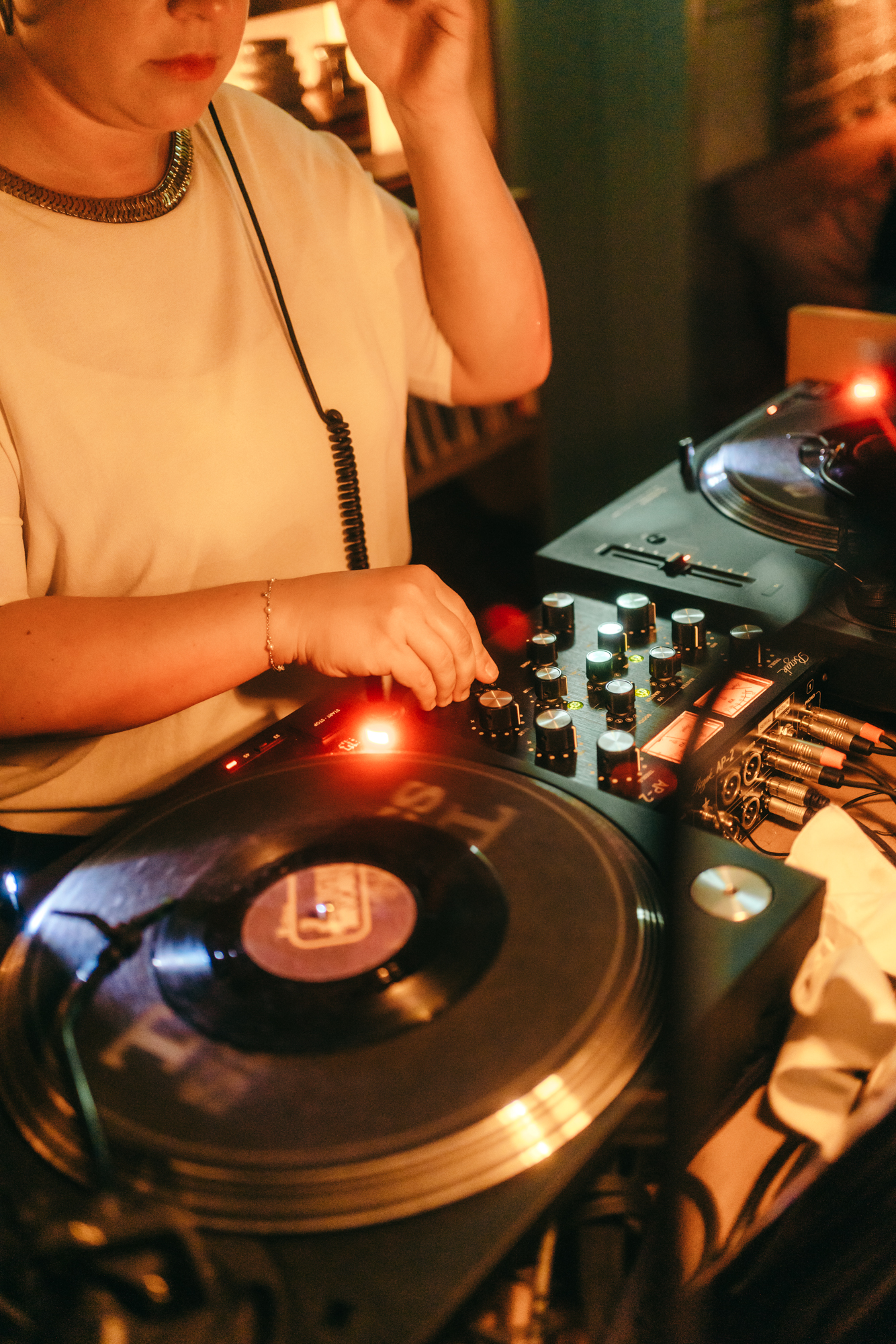 A close-up of a DJ’s hands adjusting knobs on a turntable, illuminated by warm light, with vinyl records and cables in view.